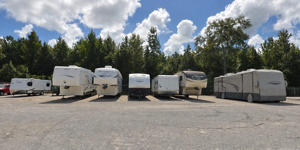 Travel trailers, fifth-wheels and a covered motorhome parked in a row on the gravel lot at Patriot Outdoor Storage, pine treeline behind